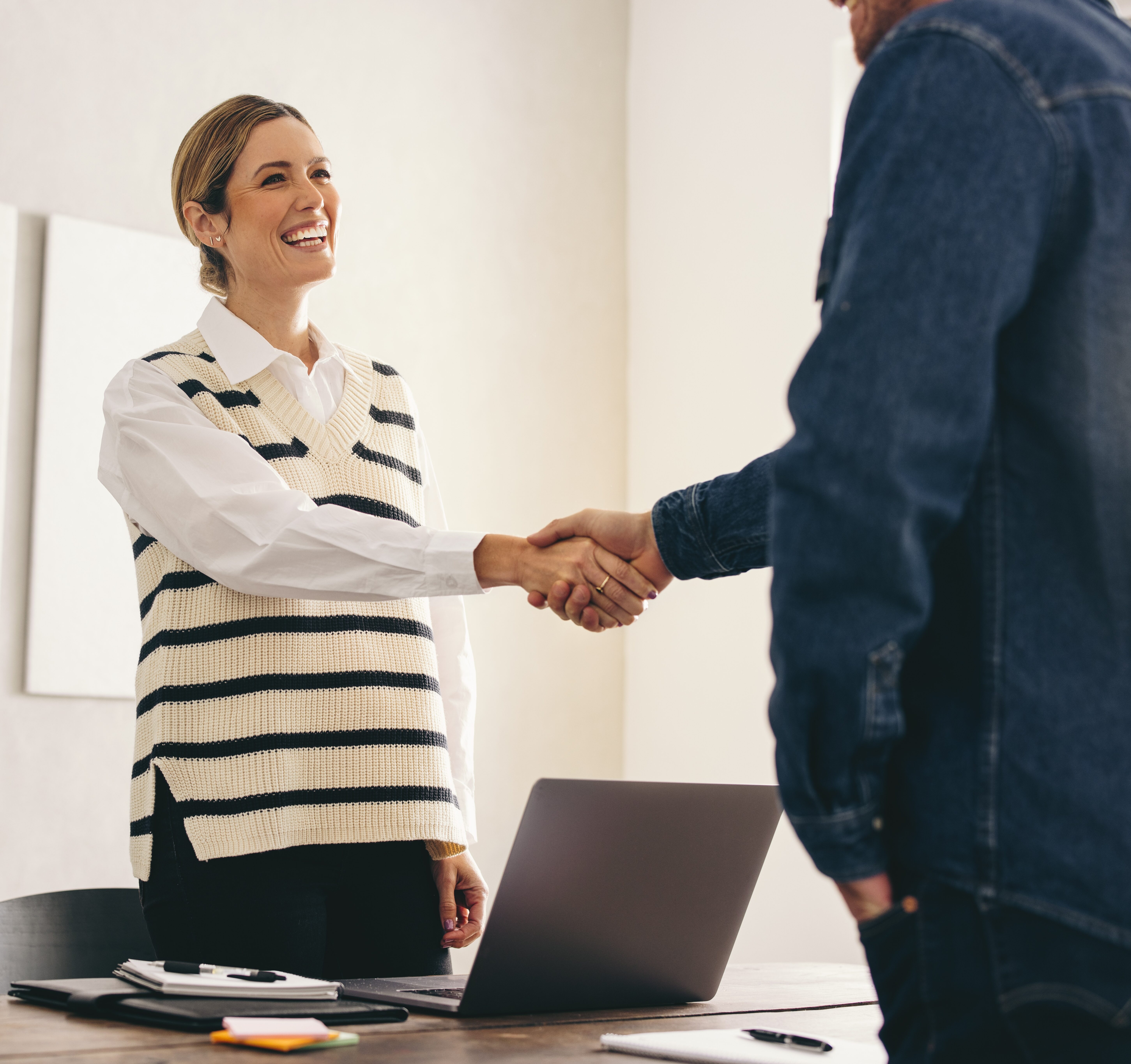 A woman standing behind a desk shakes hands with a man in an office setting, both appearing engaged and professional as they discuss group health plan administration.