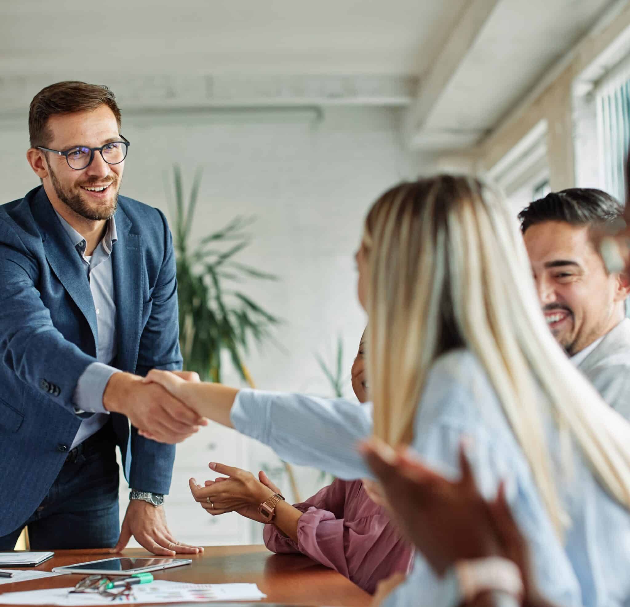 A man in a blue blazer leans over a conference table to shake hands with a seated woman during a business meeting at Providence Risk Insurance Services, while colleagues look on and applaud their partnership.