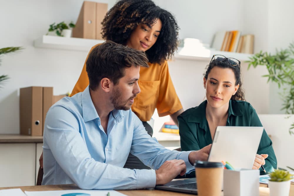Three people in a modern office work together at a desk, looking at a laptop screen as they discuss best practices for group health plans. One man sits and gestures toward the screen while two women observe.