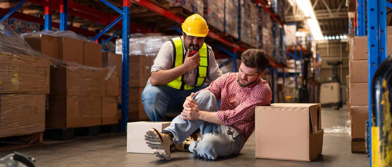 A worker sits on the warehouse floor holding his injured leg while another, in a safety vest and helmet, makes a call for assistance—highlighting the importance of group health plans in workplace safety. Boxes are stacked on shelves around them.